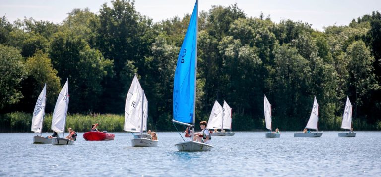 Meerdere zeilboten varen in een wedstrijdveld met een rode begeleidingsboot.