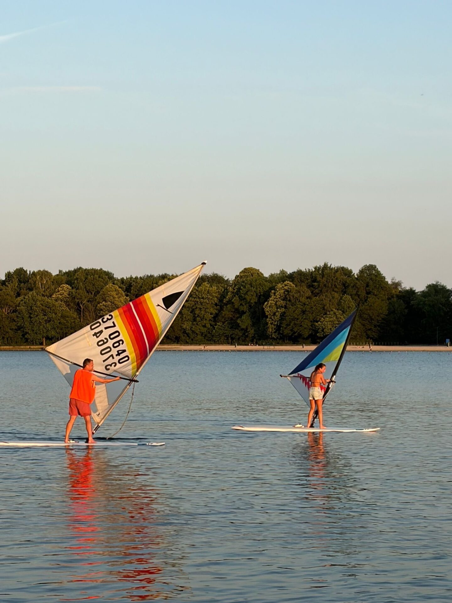 Twee windsurfers staan met kleurrijke zeilen op surfplanken bij avondlicht op de Oosterplas.